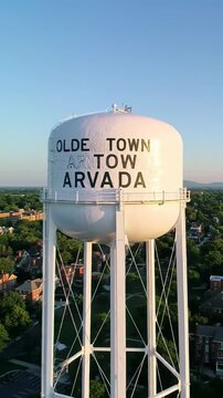 Aerial drone video unveils 'olde town arvada' water tower, suburban homes, streets, hazy mountains at golden hour. Nostalgic town atmosphere