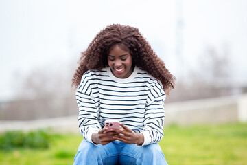 African American woman with curly long hair smiling while using smartphone outdoors