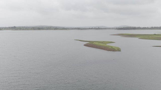 Aerial view of Chacha Kota island, Banswara, Rajasthan during monsoon, showing the lush green island surrounded by water and distant hills, with clouds hovering above.