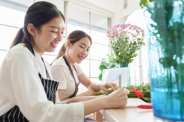 Two Asian women florists smiling while preparing flower stems and discussing business with laptop in floral shop.