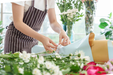 Asian woman florist wrapping bouquet with white ribbon on table, preparing floral decoration for customer order.