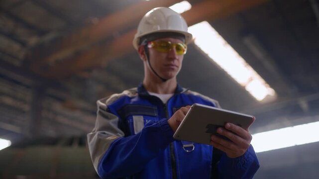 Male engineer in shipyard wearing safety gear and using tablet in industrial setting.