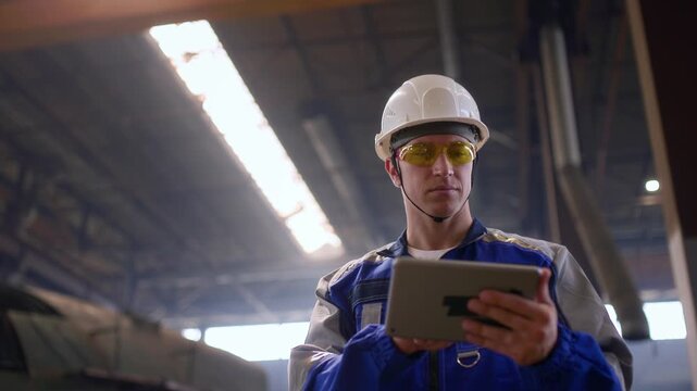 Male engineer in shipyard wearing safety gear and using tablet in industrial setting.