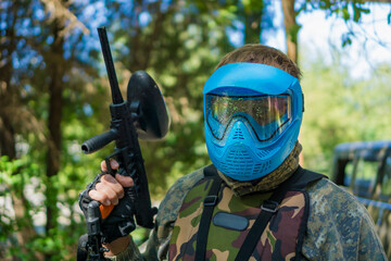Close-up of a paintball player in camouflage holding a marker gun, wearing a protective mask splattered with colorful paint after a hit.