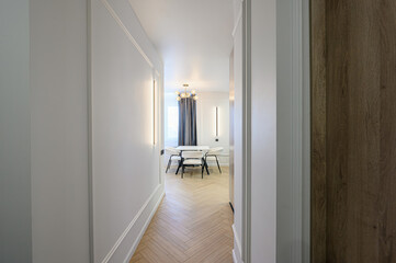 Hallway view into a modern dining area with white walls, light wood floors, and a table with white chairs. Linear wall lights provide soft illumination