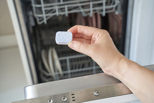 Caucasian female hand holding dishwasher tablet in kitchen with open dishwasher.