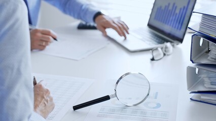 Professional colleagues reviewing financial reports, examining graphs and documents with magnifying...