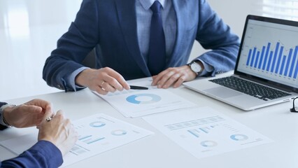 Business colleagues or partners are discussing financial data on a laptop and printed documents while analyzing charts and market trends at office table. Businesspeople and finance concept