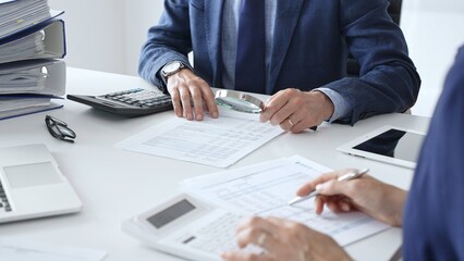 Close up of male financial analysts with female colleague examining documents, using magnifying glass and calculator at workplace in office. Audit and taxes in business