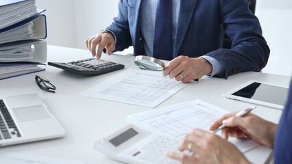 Male financial analysts with female colleague examining documents, using magnifying glass and calculator at workplace in office. Audit and taxes in business