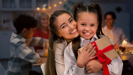 Happy young mother hugging her cute daughter with gift box during festive dinner at home. Extended multi generation family celebrating holiday together, enjoying party indoors