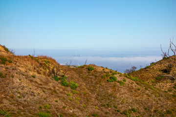 A high-angle panoramic view shows the Los Angeles urban landscape and a distant downtown skyline partially hidden by a thick marine layer of fog, as seen from a dry hiking trail in the Pacific Palisad