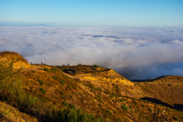 A wide-angle, high-angle view captures the scenic Pacific Palisades coastline from a dry hillside trail, with a low-lying marine layer of fog rolling over the Pacific Ocean below a clear blue sky in S
