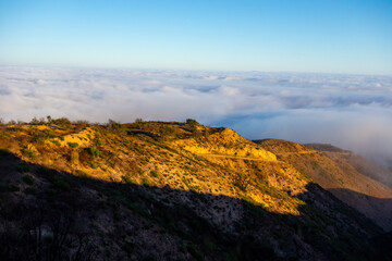 A wide-angle, high-angle view captures the scenic Pacific Palisades coastline from a dry hillside trail, with a low-lying marine layer of fog rolling over the Pacific Ocean below a clear blue sky in S