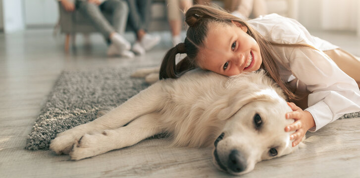 Adorable little girl lying on floor with golden retriever dog, hugging and cuddling with pet in living room, her relatives resting on sofa at home. Family times concept