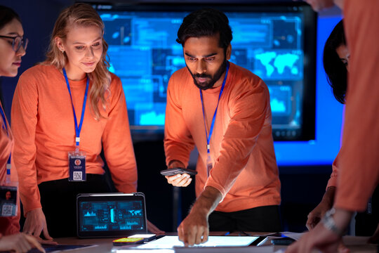 A team of data analysts in a modern control room works together on a project. The diverse group collaborates, reviewing charts, graphs, and maps on digital screens and tablets.