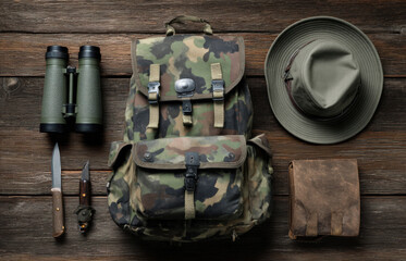 Camouflage backpack with outdoor gear including binoculars, knives, and a hat, arranged on a rustic wooden surface, showcasing adventure and exploration essentials
