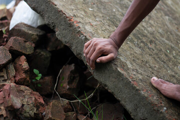 A persons hand grips a large concrete slab amidst rubble