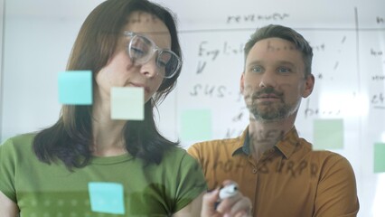 Businesswoman wearing glasses, writing on a glass board and organizing sticky notes, collaborates with a businessman observing her strategy planning in a bright office setting
