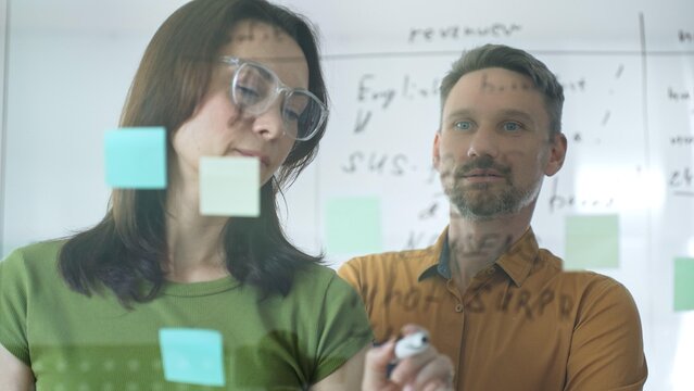 Businesswoman wearing glasses, writing on a glass board and organizing sticky notes, collaborates with a businessman observing her strategy planning in a bright office setting