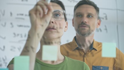 Obraz premium Businesswoman wearing glasses, writing on a glass board and organizing sticky notes, collaborates with adult businessman observing her strategy planning in a bright office