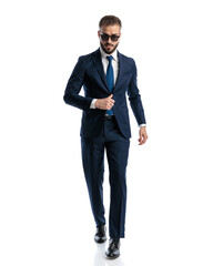 cool young man in navy blue suit with sunglasses adjusting suit and walking