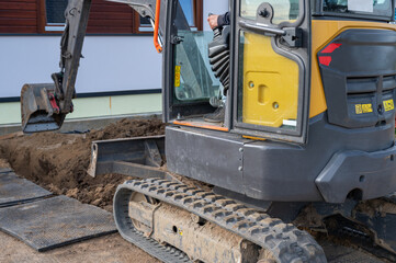 Tracked mini excavator digging trench beside residential building wall.