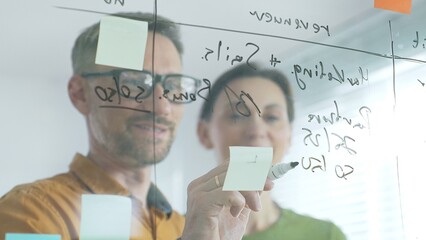 Business team of professionals brainstorming near glass board, using sticky notes for strategic planning, writing calculations, sharing innovative ideas during collaborative work