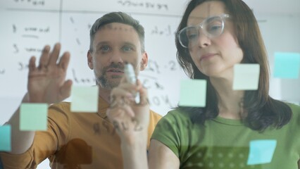 Businesswoman wearing glasses, writing on a glass board and organizing sticky notes, collaborates with a businessman observing her strategy planning in a bright office setting