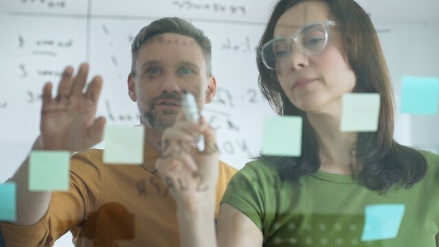 Businesswoman wearing glasses, writing on a glass board and organizing sticky notes, collaborates with a businessman observing her strategy planning in a bright office setting