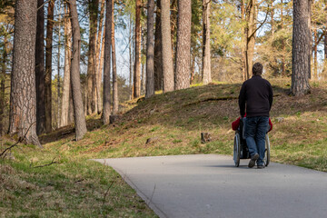 View from back to a woman in wheelchair and a man pushing her in forest park at paved road. Woman with a disability in wheelchair and her personal assistant are walking together