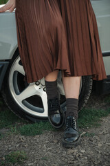 Fototapeta premium lose-up of woman’s legs in pleated skirt and black shoes walking near a car wheel on a countryside road, casual fashion and lifestyle detail.