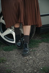 lose-up of woman&rsquo;s legs in pleated skirt and black shoes walking near a car wheel on a countryside road, casual fashion and lifestyle detail.