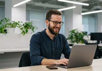 A smiling bearded man wearing glasses works diligently on his laptop in a modern well lit office environment with lush greenery