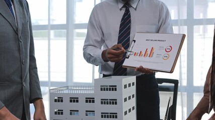 Professional architect wearing business attire presenting building project to investor, using clipboard with charts and small scale architectural model during collaborative meeting - Powered by Adobe