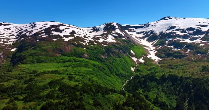 Flying closer to the beautiful green mountains covered with snow on top. Sunny scenery of the amazing rocks in the wilderness of Alaska, USA.