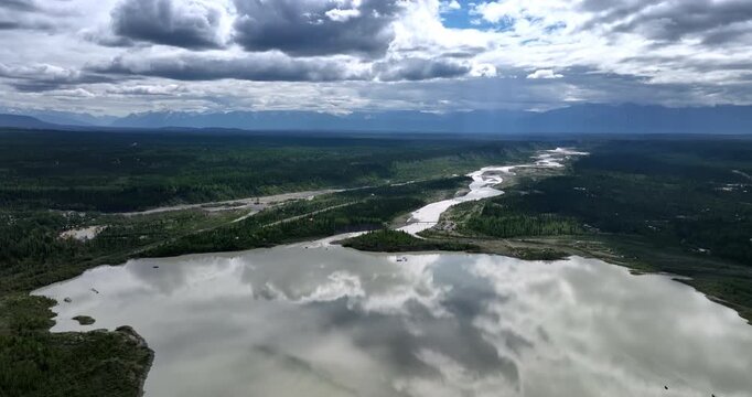 Grey clouds reflect in the river. Evergreen pine tree woods grow around the waterscape. Aerial view. Alaska, USA.