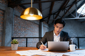 Focused Asian businessman working on laptop at desk with blueprints and coffee