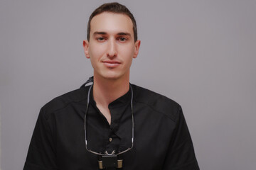 Smiling doctor in black uniform. A healthcare dentist, gynecologist, veterinarian. A specialist in medicine. Binocular glasses are hanging around the doctor's neck. The doctor on a gray background. 