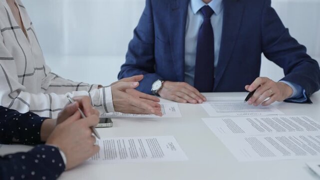 Business professionals analyzing contract details, gesturing with pens while discussing legal terms during collaborative meeting, focused on negotiation process