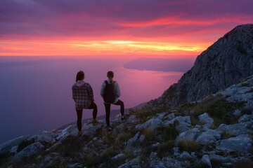 Fototapeta premium Two women hikers with backpacks standing on rocky mountain peak during sunset in Italy, overlooking the sea with dramatic colorful pink sky. Outdoor travel adventure. Sporty girls on mountain at dusk