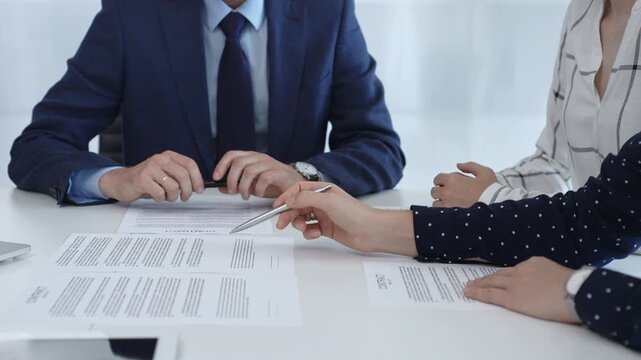 Business professionals analyzing contract details, gesturing with pens while discussing legal terms during collaborative meeting, focused on negotiation process