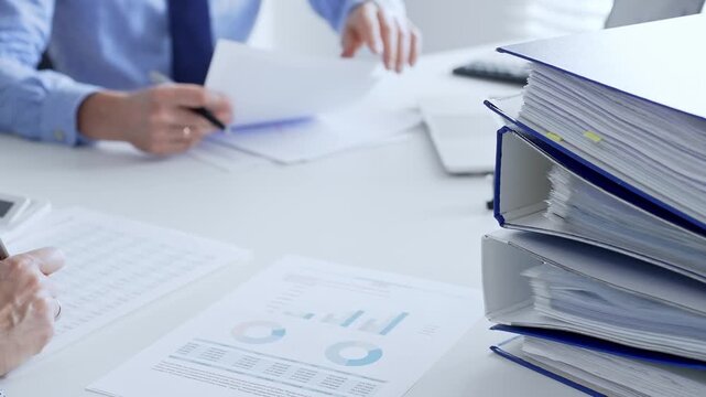 Corporate professionals reviewing financial documents, working collaboratively at white desk with blue folder stack, highlighting teamwork and business productivity. Audit and taxes in business