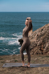Athletic woman in form-fitting activewear stretches on yoga mat by the ocean, showcasing flexibility and strength against a scenic coastal backdrop