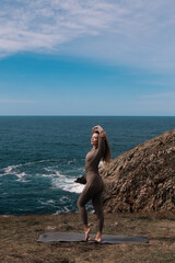 Athletic woman in form-fitting activewear stretches on yoga mat by the ocean, showcasing flexibility and strength against a scenic coastal backdrop with blue skies