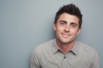 Obraz premium Confident young man with short dark hair smiling wearing casual grey shirt against plain grey background.