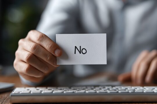 A Close-Up of a Hand Holding a Card with the Word 'No', Symbolizing Refusal or Rejection in a Professional Setting with a Keyboard in the Background