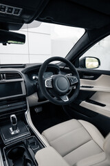Modern car interior with light beige leather. Driver's seat of right-hand drive luxury car: steering wheel, dashboard and center console of a new vehicle parked near warehouse.