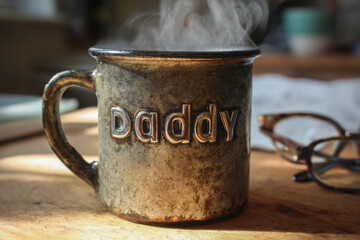 Rustic metal mug with steaming hot drink and embossed lettering placed on wooden table near reading glasses in cozy home setting