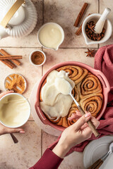 Female hands pouring glaze over freshly baked cinnabon buns in ceramic baking dish, beige tile background. Making cinnamon rolls process, top view